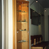 Glasvitrine aus Buche im Vorraum einer Praxis. Tür und Böden aus Glas. Die Vitrine steht vor einem Wandvorsprung